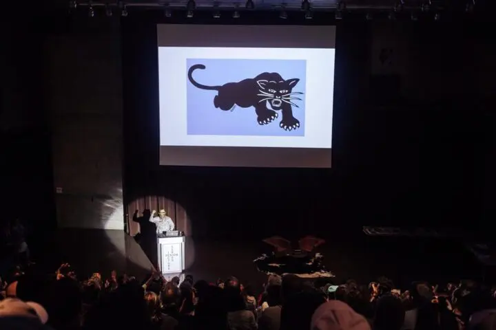 Audience watching a presentation by Emory Douglas of a black panther graphic on a large...
