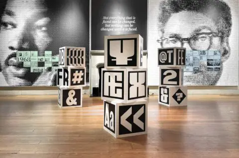 Art exhibition with typographic blocks and portraits on walls, featuring a quote about ...