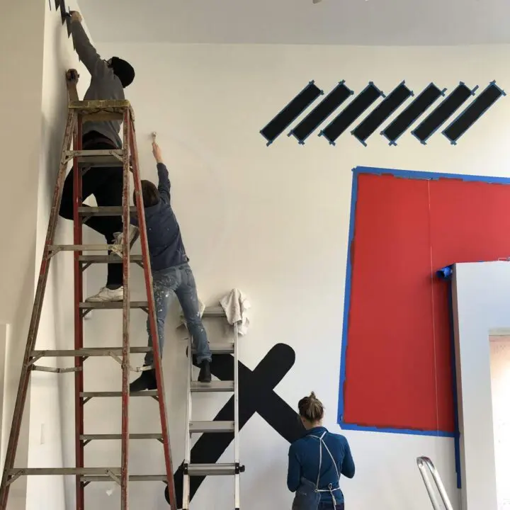 Civilization painting a Na Kim wall mural with geometric patterns using ladders in a br...