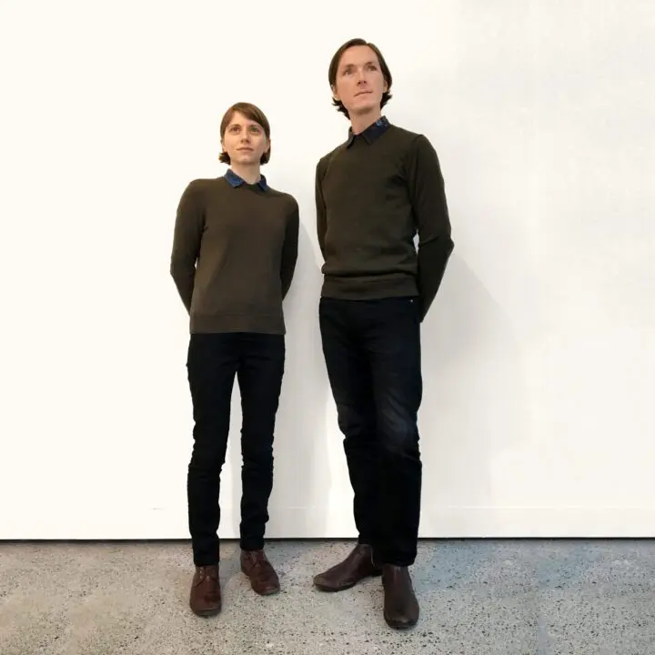 Molly Derse and Shaun Kardinal standing against a white wall, both wearing dark sweater...