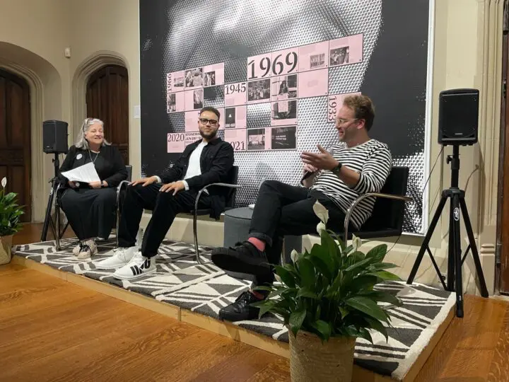 Three people engage in a discussion panel against a backdrop with historical dates, sea...