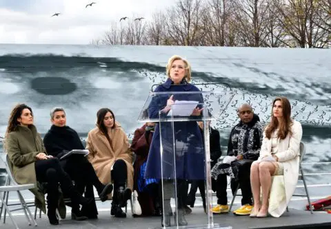 A woman speaks at a podium outdoors, surrounded by seated listeners on a stage, with bi...
