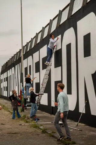 People painting a large mural on a tall wall, using ladders and teamwork for artistic s...