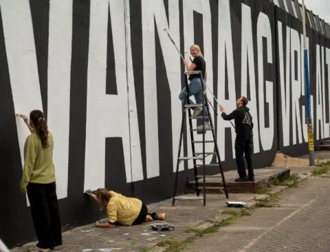 Artists painting large black and white text mural on a wall, using ladders and brushes ...