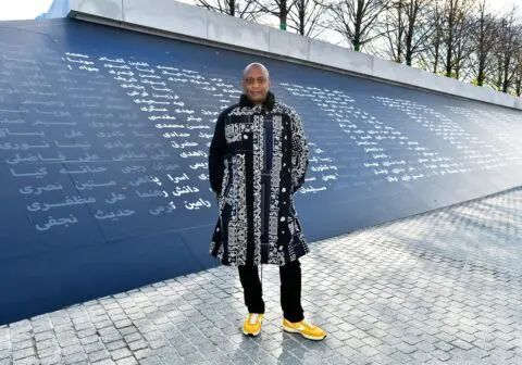 Man in patterned coat and yellow shoes stands in front of a wall with text in various l...