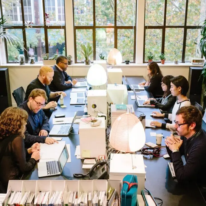 Open-plan office with diverse team collaborating at laptops, surrounded by plants and c...
