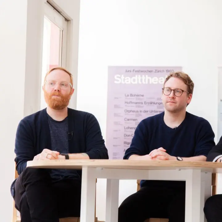 Three men sitting at a table in a modern room with theater posters behind them.