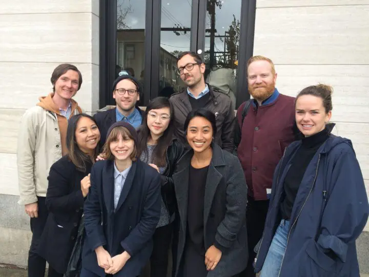 A group of nine people smiling outside a building, wearing casual and semi-formal outfi...