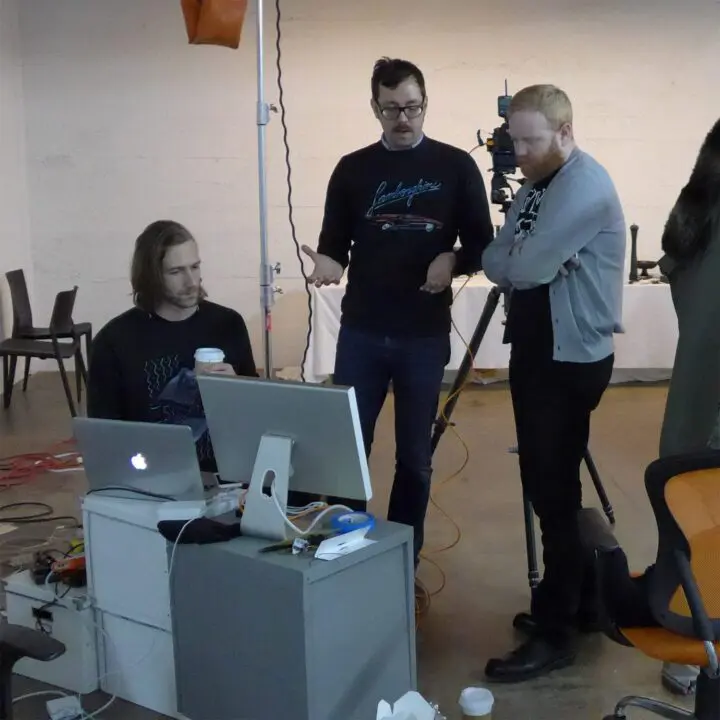 Three men collaborate on a project in a studio; one seated, two standing by a computer ...