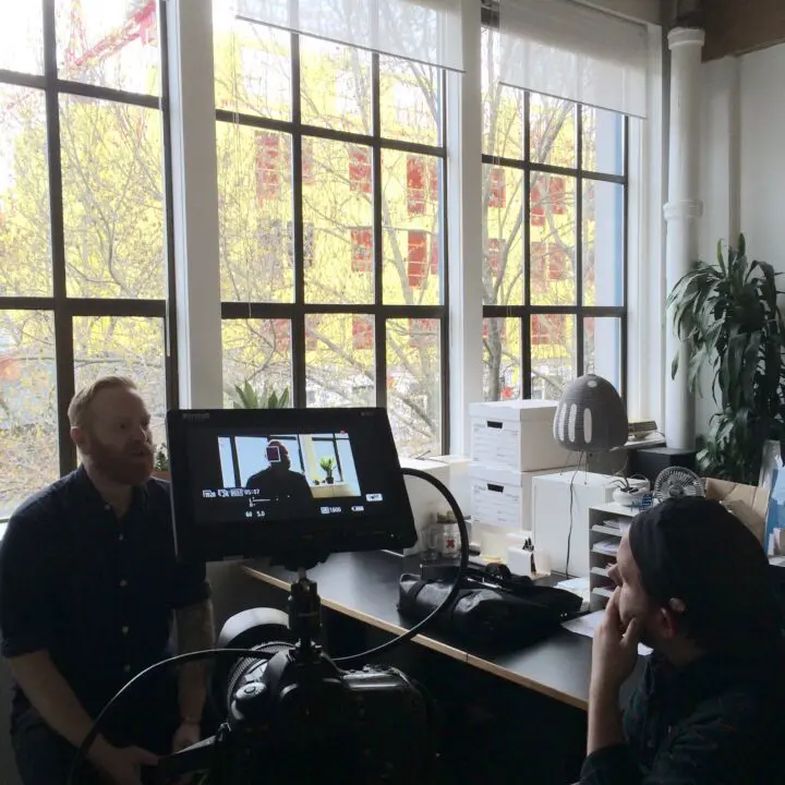 Man being filmed in an office setting with large windows, camera and monitor visible; c...