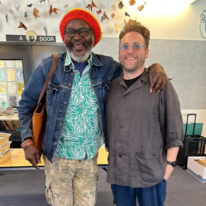 Michael Ellsworth and Saki Mafundikwa smiling and posing together indoors, one in a den...
