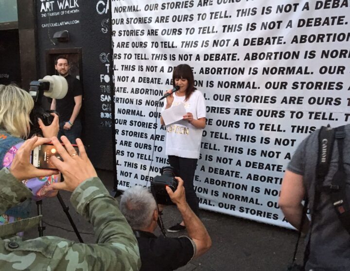 Person speaking at an event with Abortion is Normal banner, surrounded by photographers...