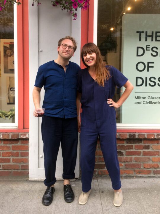 Two people smiling and posing outside a building with red brick and a design poster in ...