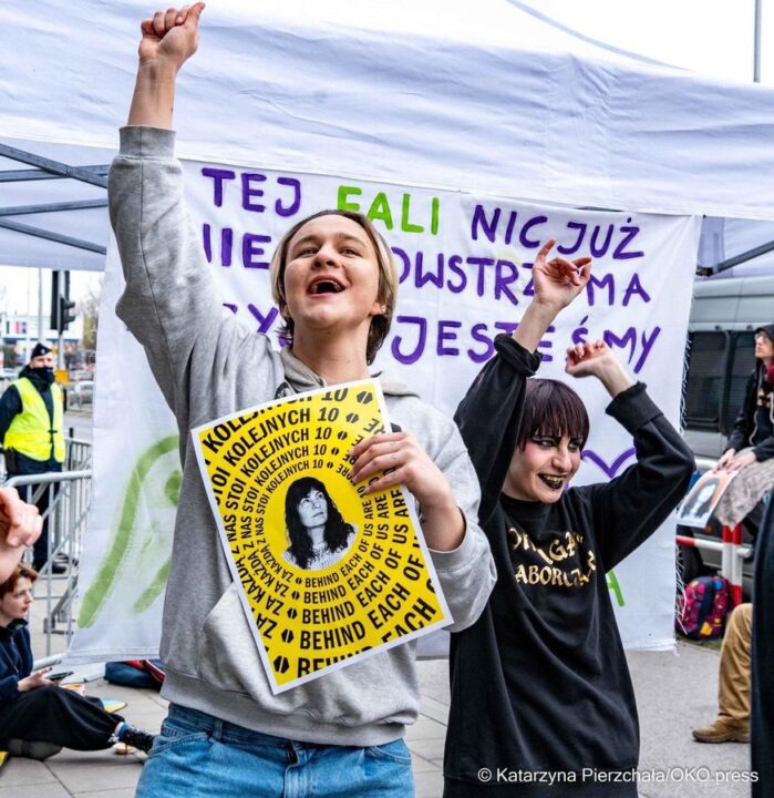 People at a protest, smiling and raising fists, holding a sign with text in Polish and ...