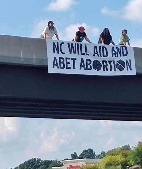 Protesters on a bridge display sign reading NC Will Aid and Abet Abortion with blue sky...