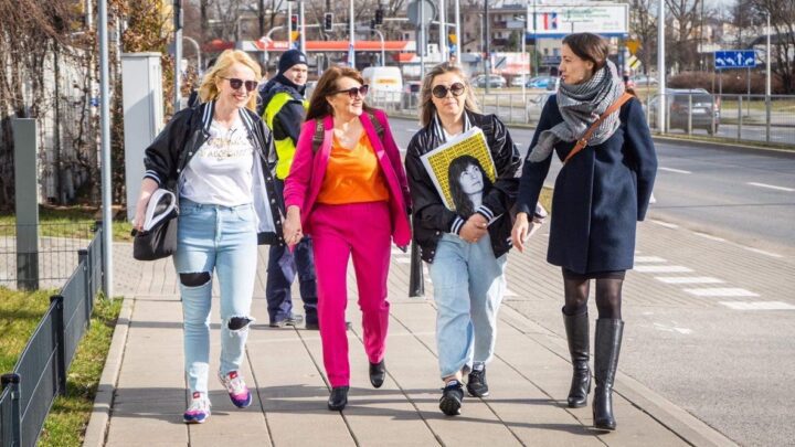 Four women walking confidently on a city sidewalk, enjoying a sunny day, with one holdi...