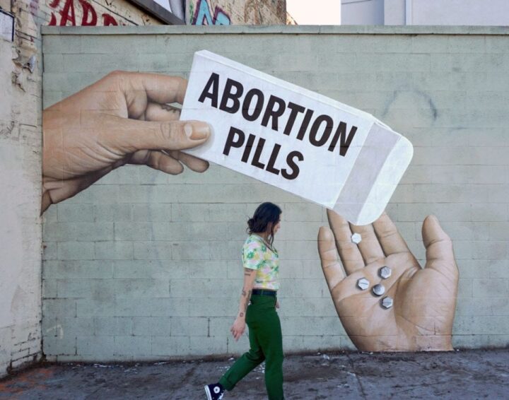 Street mural depicting large hands offering abortion pills, with a person walking by. U...