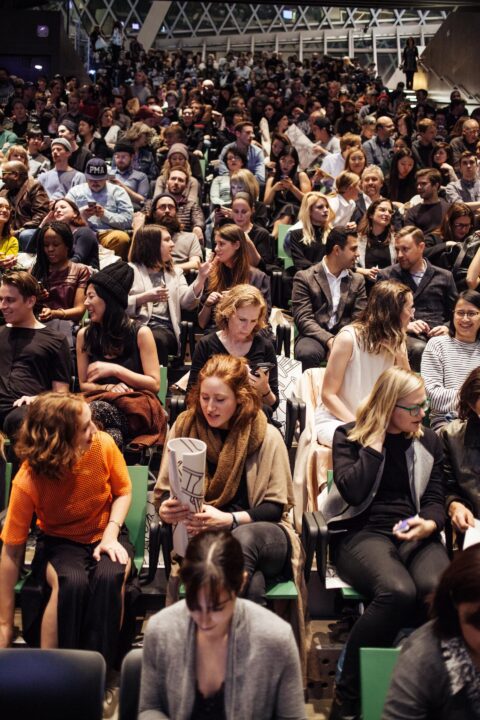 Audience eagerly awaits Design Lecture Series start in packed auditorium, diverse crowd chatting and engaging.