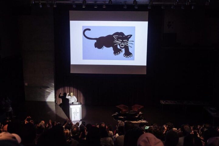 Audience watching a presentation by Emory Douglas of a black panther graphic on a large screen in a dimly lit auditorium.