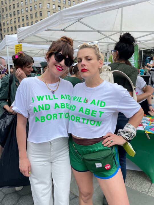 Two women at a rally wearing white t-shirts with I WILL AID AND ABET ABORTION text, pro...