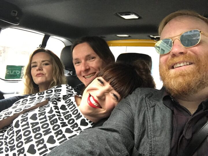 Group of friends smiling in a car, one woman wearing an abortion rights-themed outfit, ...
