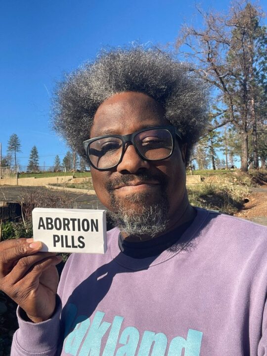 Man outdoors holding a box labeled Abortion Pills, promoting reproductive rights under ...