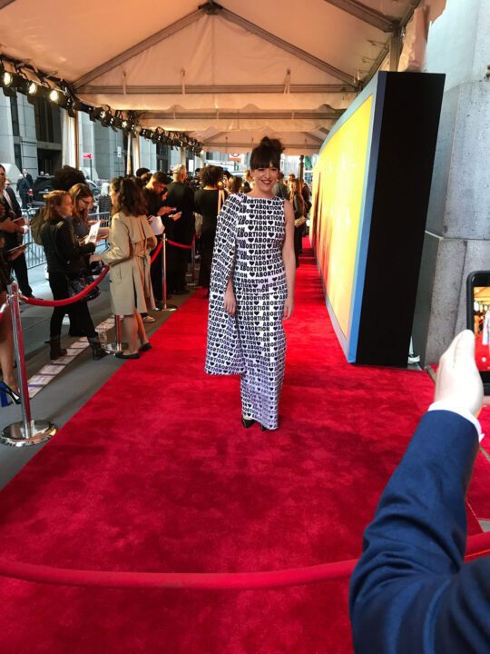 Person in bold text dress on red carpet, surrounded by photographers at a high-profile ...