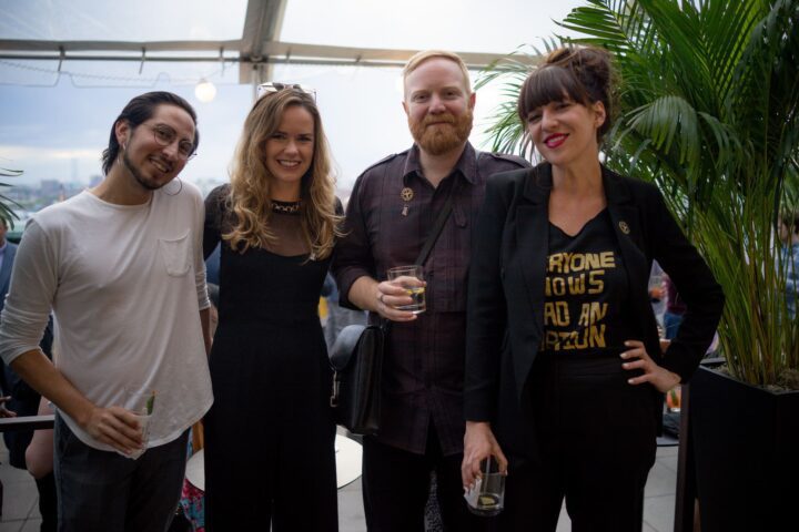 Group of four friends enjoying drinks at a rooftop event, smiling for the camera, surro...