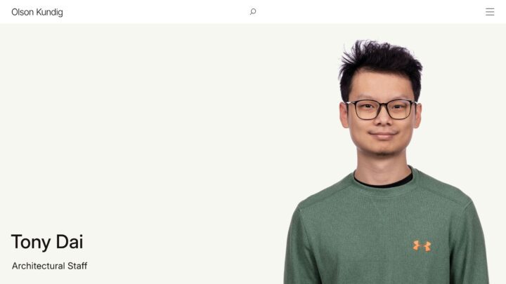 Person smiling in front of a light background wearing glasses and a green sweater, show...