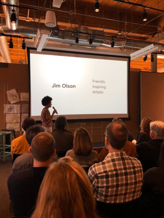 Raya Leary presenting our website design to the Olson Kundig staff at their Seattle office with a slide showing Jim Olson - Friendly, Inspiring, Artistic in a modern conference setting.