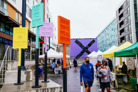 Vibrant city street with colorful signs promoting equality and human rights, surrounded by market stalls and modern buildings.