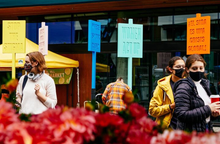 People wearing masks at a protest with colorful signs about social issues, set against ...