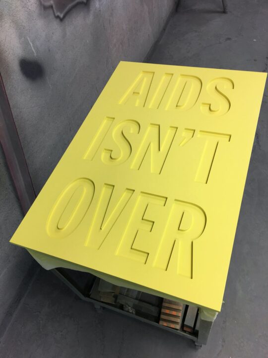 Yellow sign with embossed text AIDS ISN'T OVER highlighting the ongoing need for awaren...