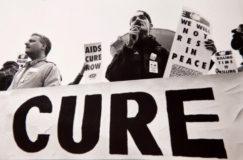 Activists at an AIDS protest holding signs for a cure. Signs read AIDS CURE NOW and WE WILL NOT REST IN PEACE.