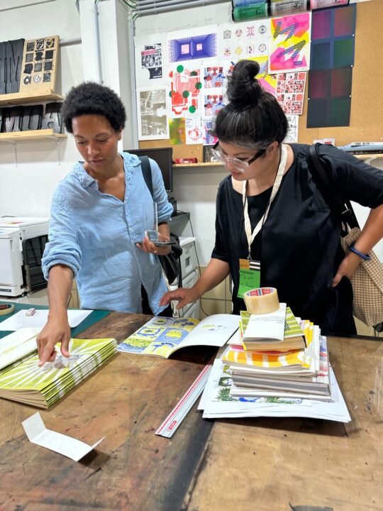 Raya Leary and Trish Chua browse colorful prints and books in an art studio, surrounded by vibrant wall designs and creative materials.