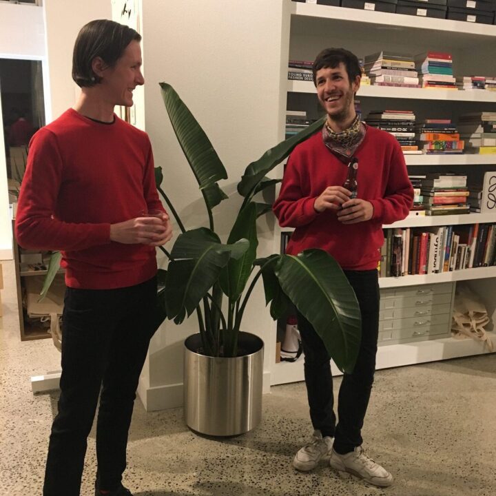 Shaun Kardinal and Oscar Viñas in matching red sweaters sharing a conversation next to a houseplant, with bookshelves in the background.
