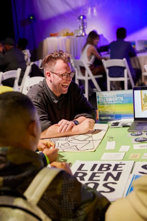 Michael Ellsworth laughing at a table for the teen design event at the Cooper Hewitt Smithsonian Design Museum with design materials and a laptop, surrounded by people at a creative networking event.