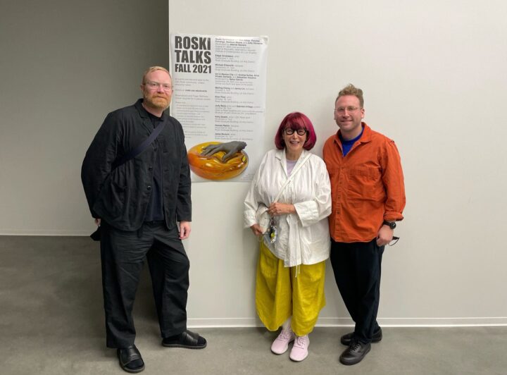 Corey Gutch, April Greiman, and Michael Ellsworth standing by a Roski Talks Fall 2021 poster, dressed in colorful outfits, in an art gallery setting.