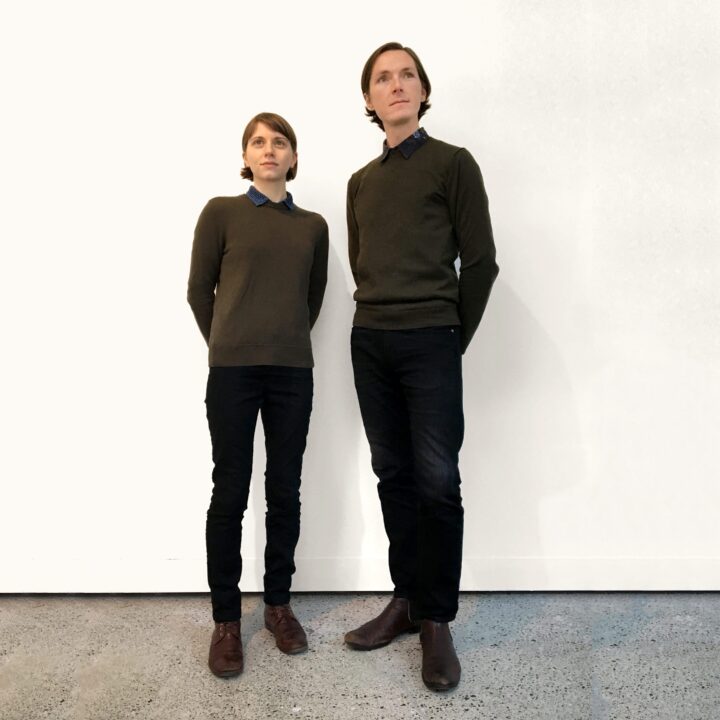 Molly Derse and Shaun Kardinal standing against a white wall, both wearing dark sweaters and shoes, looking upward.