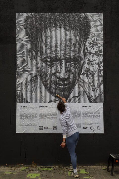 Artist painting a mural featuring a large monochrome portrait on a black wall, showcasi...