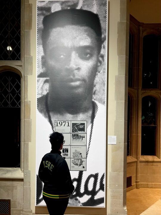 Person viewing a large, black-and-white portrait poster in a gallery, with text and ima...