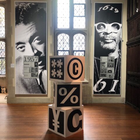 Modern art exhibit with graphic cubes and historical photo collages, displayed in a sunlit gallery with large windows.