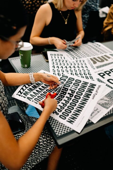 Person cutting out letters and numbers from printed sheets at a table, with scissors and a green cup nearby.