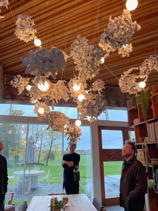 Michael Ellsworth, Corey Gutch, and Randy Bishop examine a Bocci 44 Series lighting chandelier in a bright room with large windows and green outdoor view.
