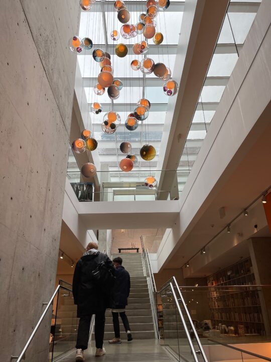 Modern atrium with colorful hanging glass art, two people ascending stairs, natural lig...