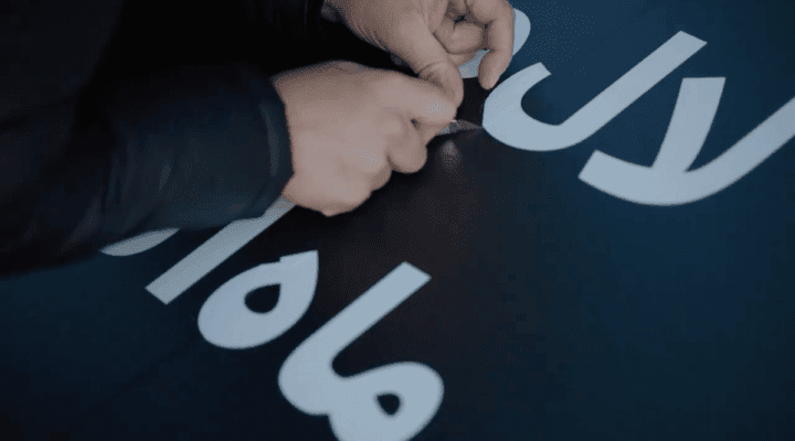 Close-up of hands applying vinyl letters on a dark surface, using a knife for precise p...