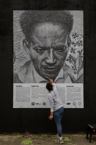 Artist painting large black and white mural on wall, depicting a serious face with text below, urban art scene.