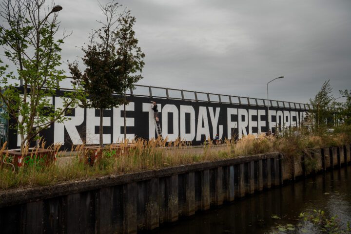 Mural with large text FREE TODAY, FREE FOREVER on an outdoor wall, amidst urban greener...