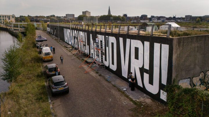 Aerial view of artists painting large white letters on a long wall beside a river in an...