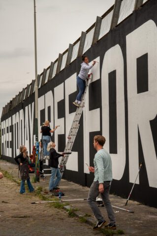 People painting a large mural on a tall wall, using ladders and teamwork for artistic street art creation.